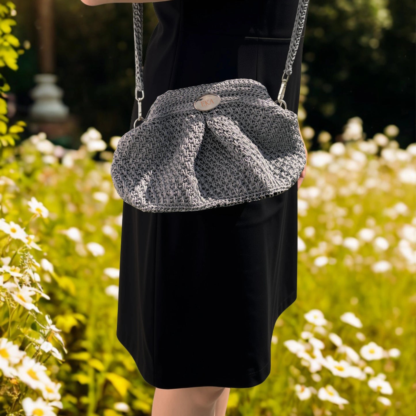 A person wearing a black dress carries a silver crocheted shoulder bag with a chain strap in a sunlit garden with white flowers.