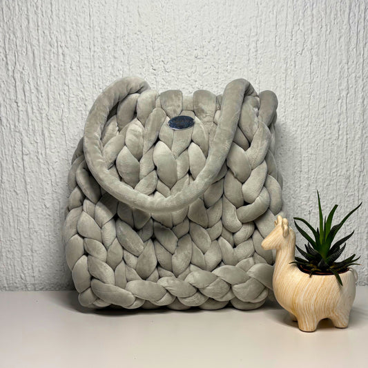 Gray braided purse with a small plant and decorative item on a light surface against a white wall.