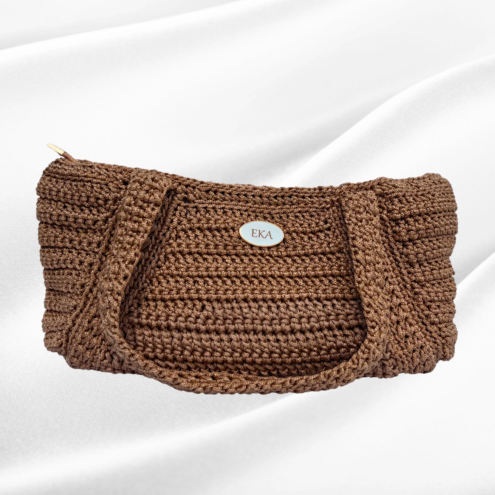 Front view of the handcrafted Elena shoulder bag by Eka in metallic bronze-gold crochet, featuring an oval gold logo plaque centered on the woven texture.