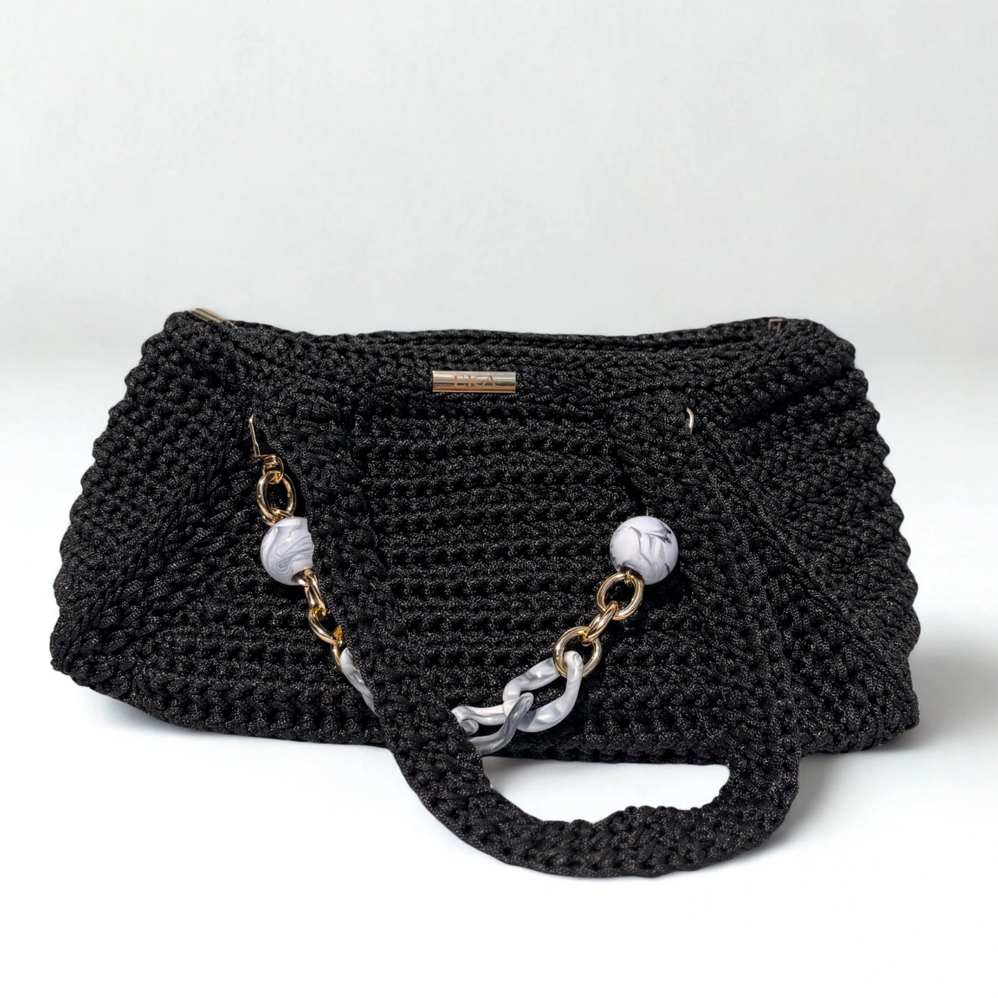 A black crocheted shoulder bag with a textured knit body, featuring a long strap adorned with white marble beads and gold-tone chain links, set against a plain light gray background.