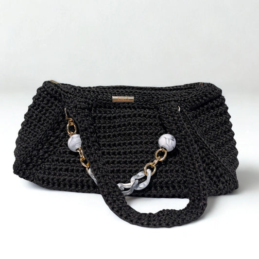 A black crocheted shoulder bag with a textured knit body, featuring a long strap adorned with white marble beads and gold-tone chain links, set against a plain light gray background.