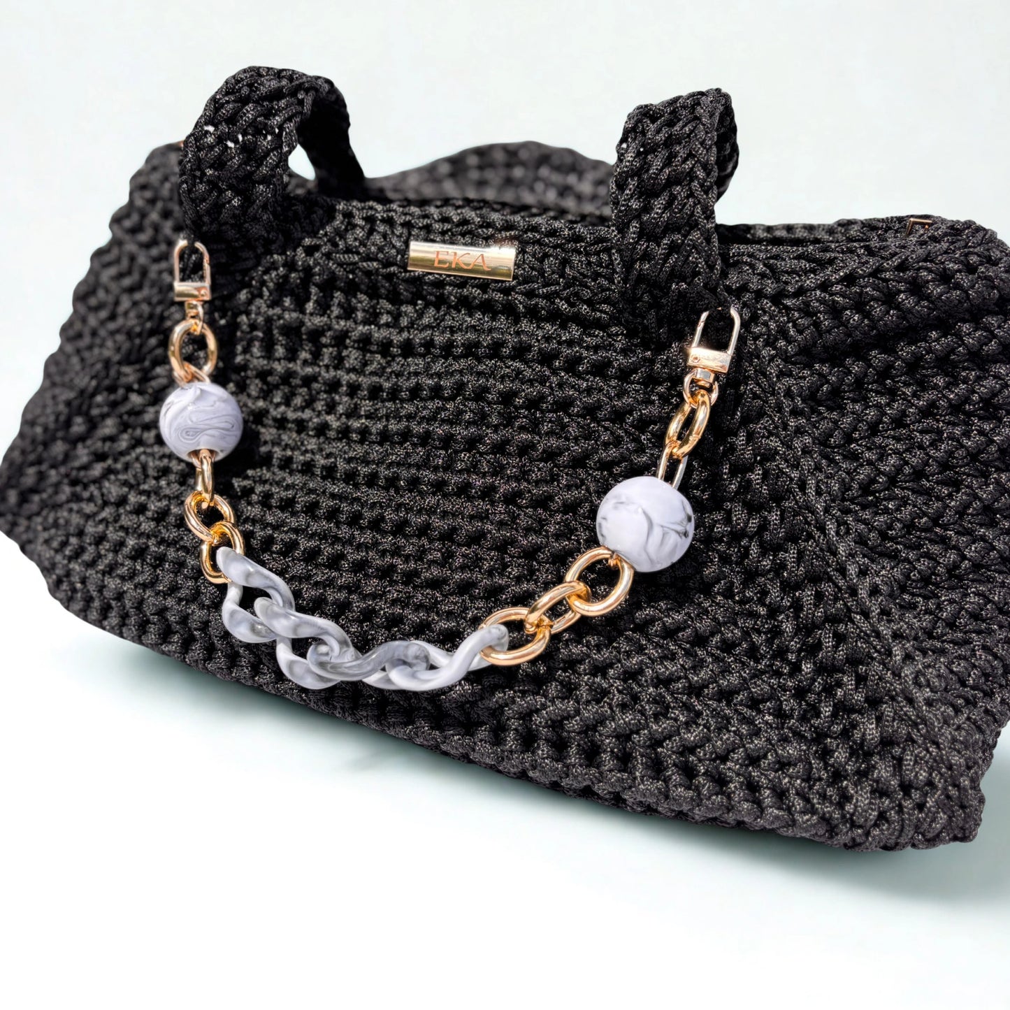 A black crocheted handbag with a decorative chain strap featuring gold-tone links, a translucent gray chain link, and two round white marble beads. A small gold metal plaque "EKA" is visible on the front.