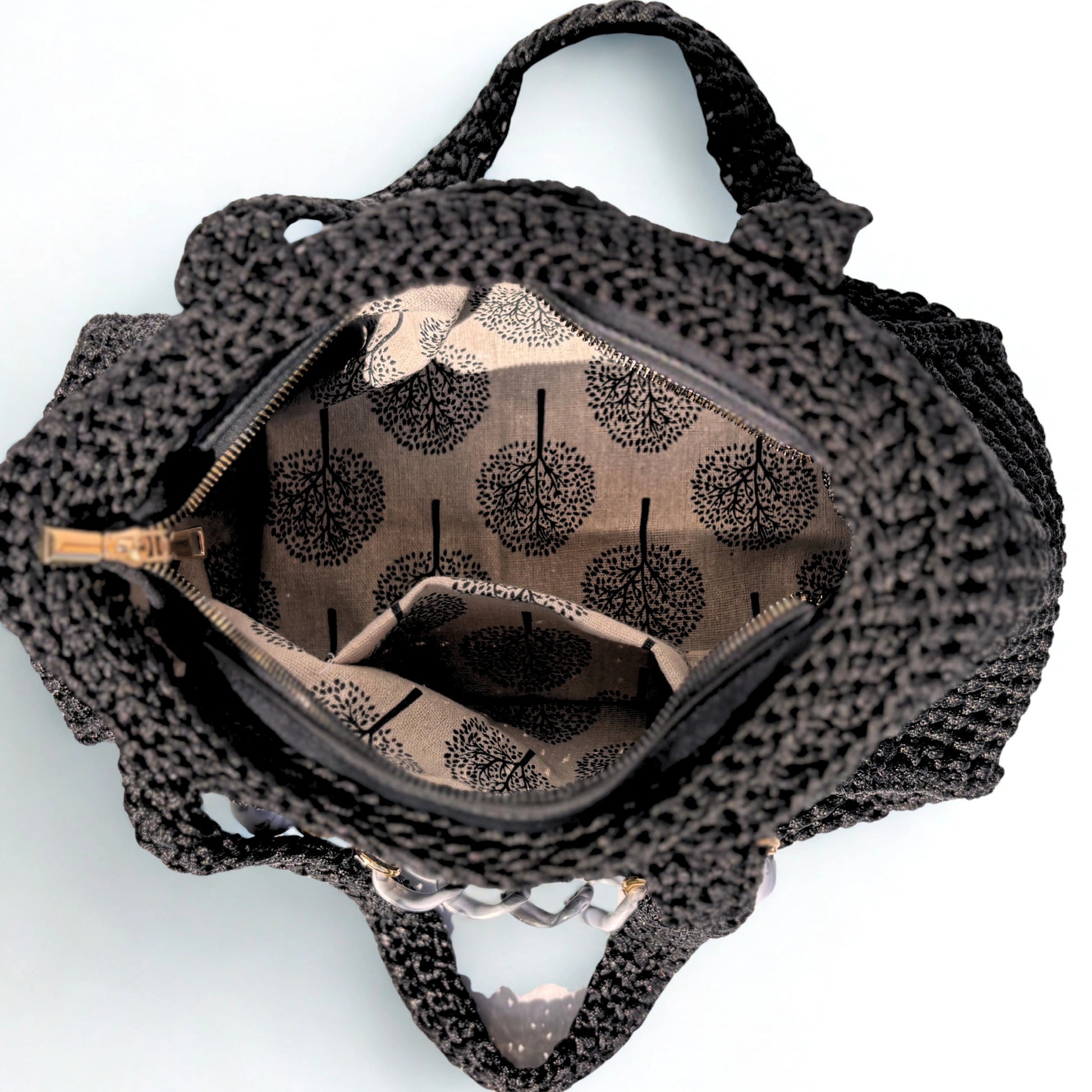 An overhead view of a black crochet handbag with a patterned brown and beige interior lining featuring a tree print, shown on a white background.