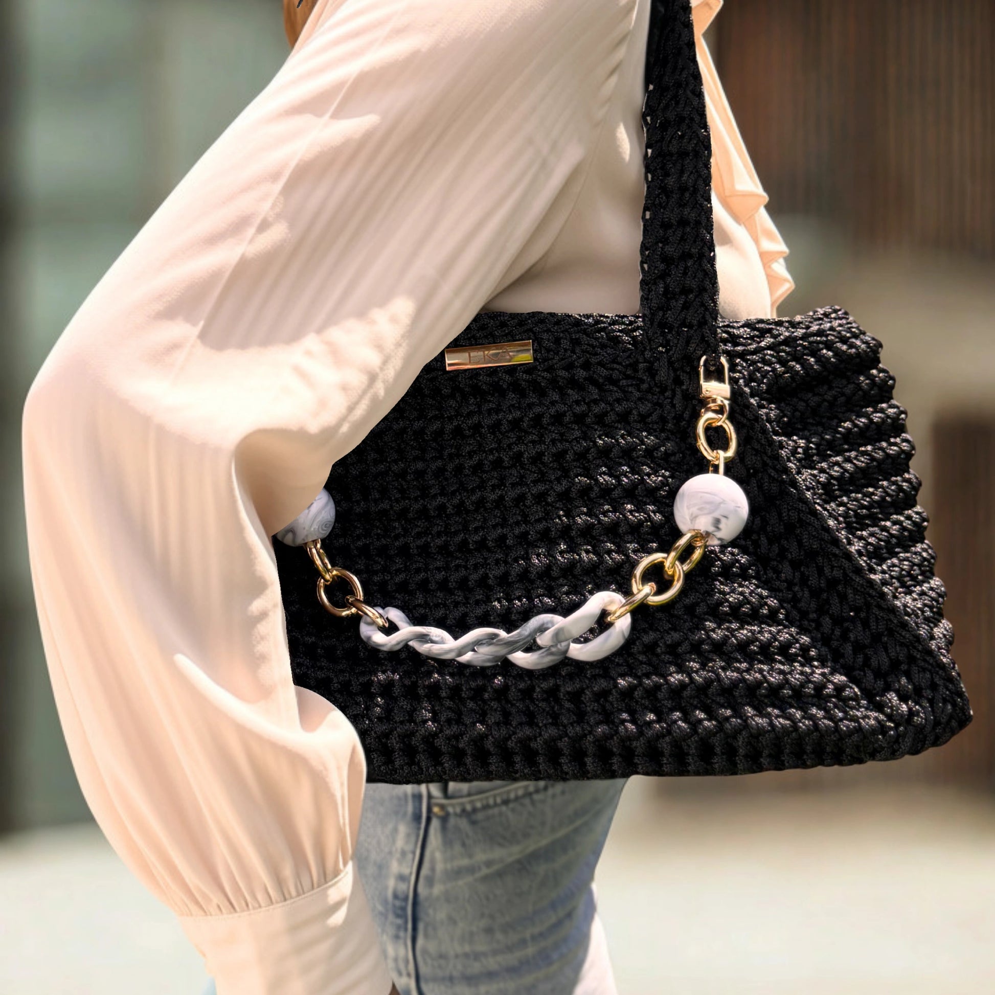 A person wearing a cream pleated blouse and light blue jeans carries a large black crocheted shoulder bag with a chunky silver chain and gold-tone hardware.