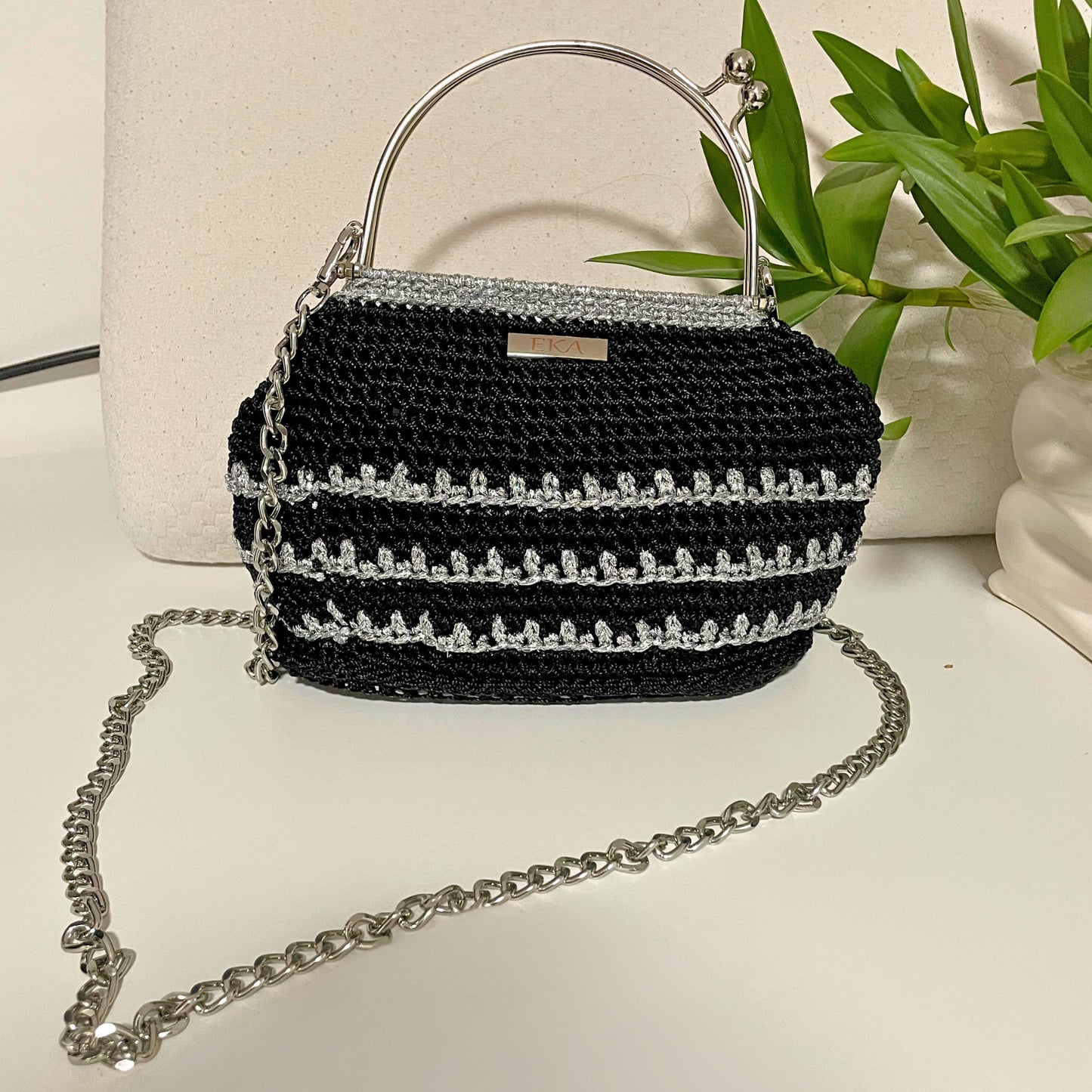 Black crochet handbag with silver chain strap on a light surface with a plant in the background