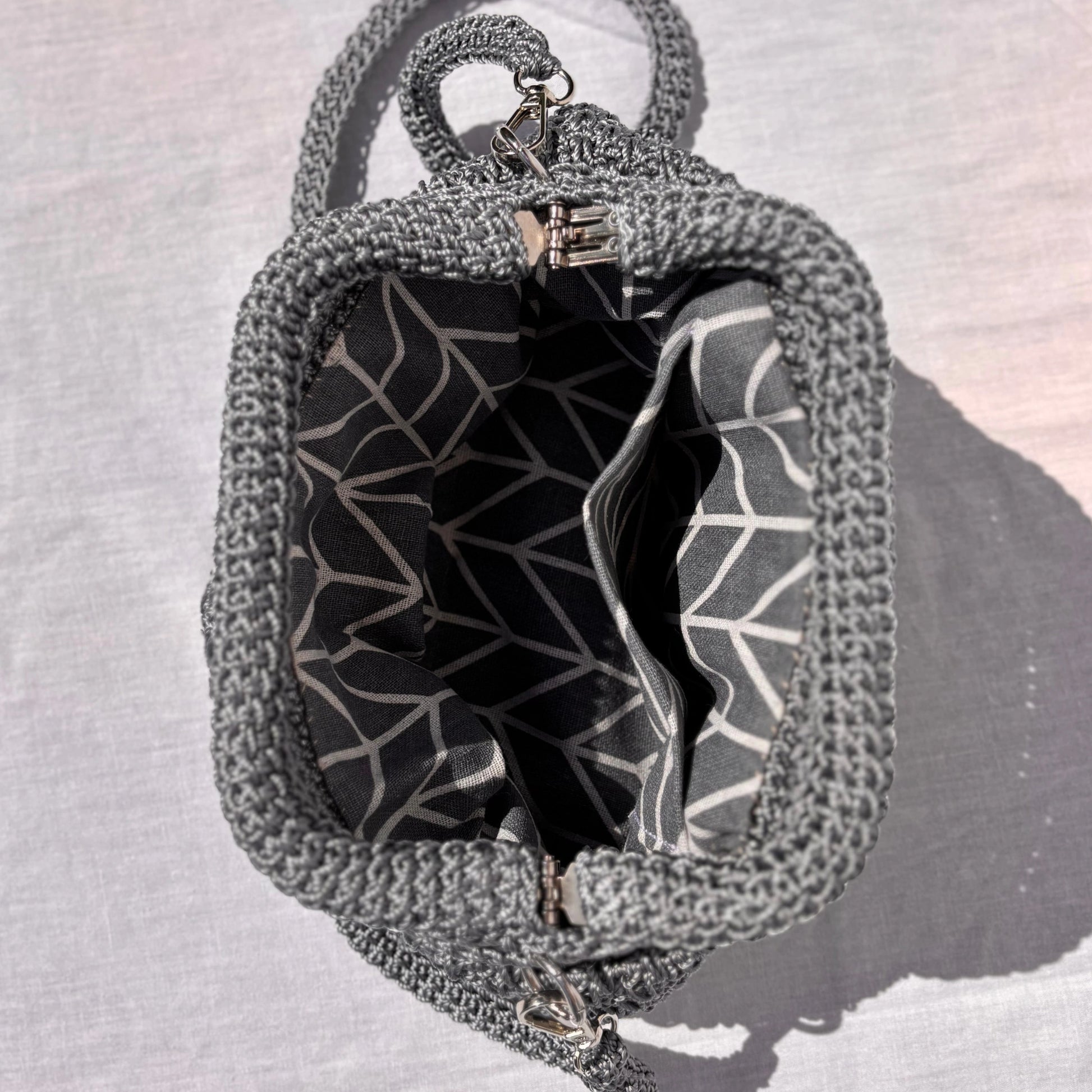 A close-up of a gray crocheted handbag viewed from above, showing a black and white geometric patterned interior lining and a silver zipper closure, set against a light gray fabric background.