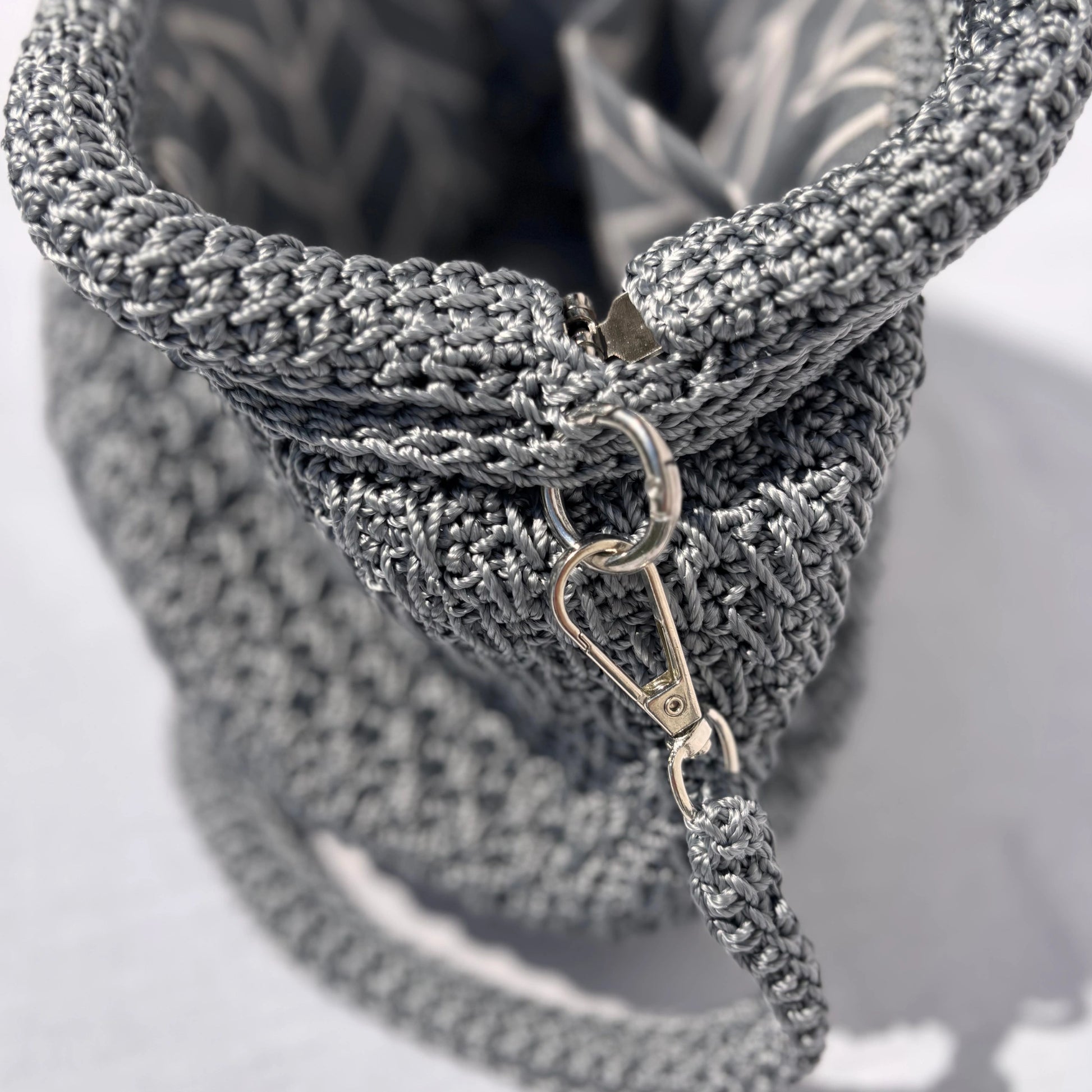 Close-up of a silver crocheted bag with a textured woven body, silver metal clasp, and matching strap on a light gray background.