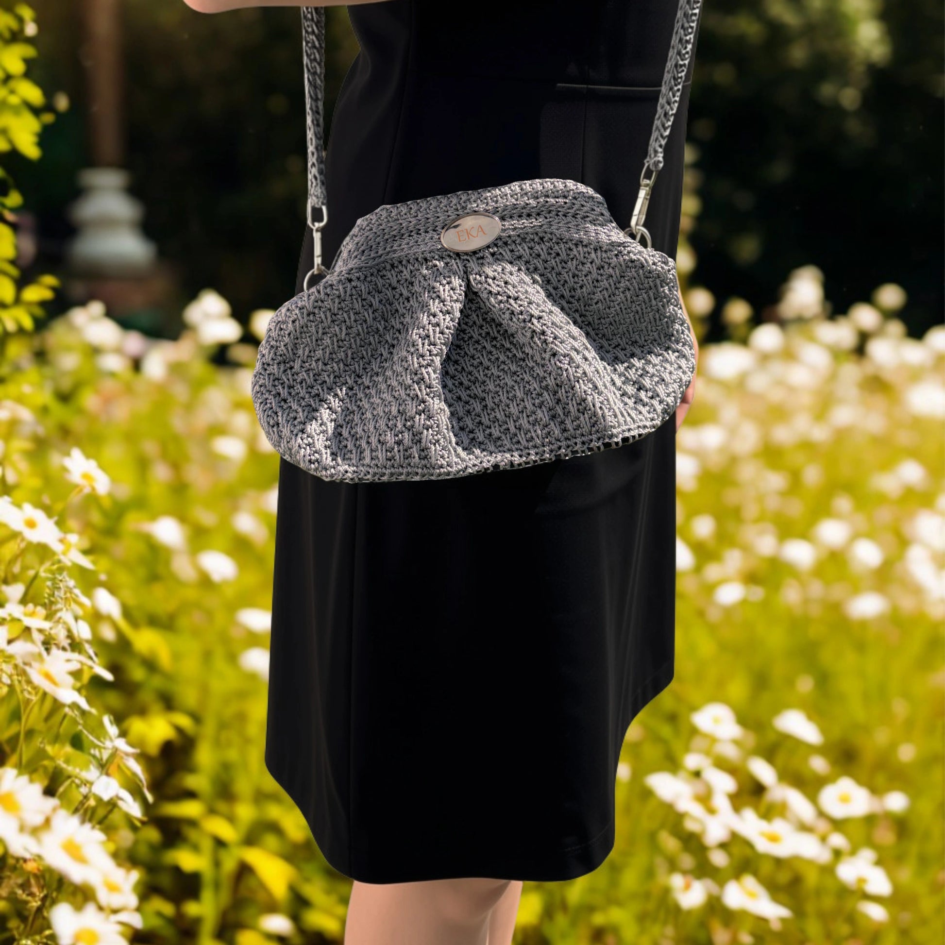 A person wearing a black dress carries a silver crocheted shoulder bag with a chain strap in a sunlit garden with white flowers.