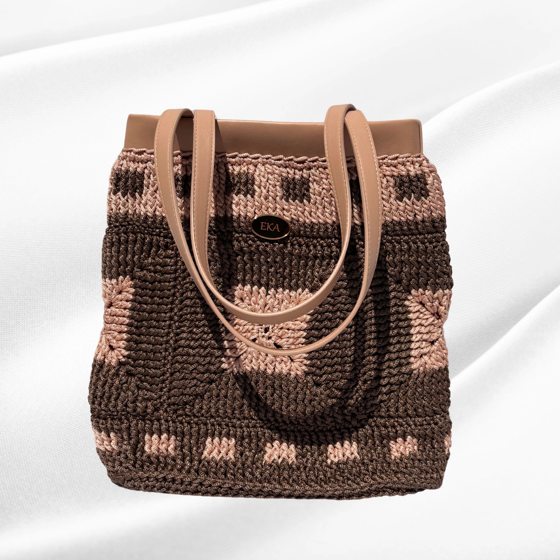 Front view of the Fiona crochet tote purse by Eka, showing the brown and blush-pink geometric granny square pattern, nude leatherette top trim, dual shoulder straps, and gold EKA emblem.