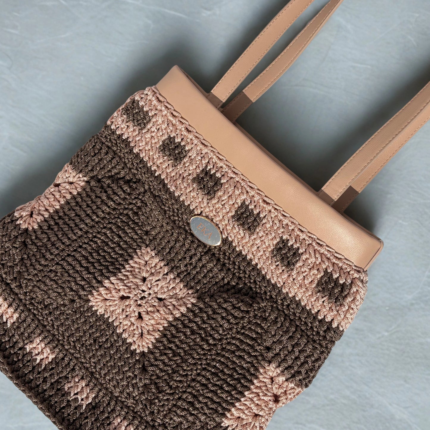 Flat-lay view of the Fiona tote bag highlighting the detailed crochet texture, nude-tan faux-leather collar, long shoulder handles, and oval metal logo plaque.