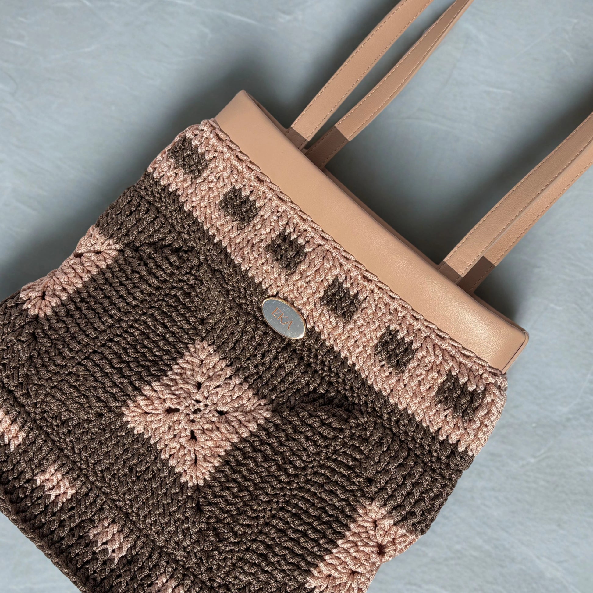Flat-lay view of the Fiona tote bag highlighting the detailed crochet texture, nude-tan faux-leather collar, long shoulder handles, and oval metal logo plaque.