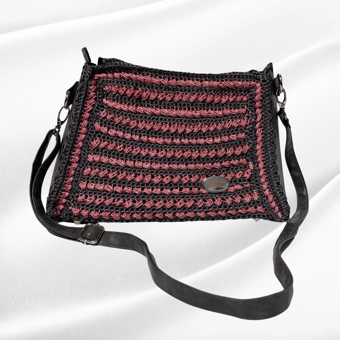 Front view of the Gloria handcrafted purse by Eka, featuring a dark charcoal grey and dusty rose pink textured crochet pattern, real leather side panels, an adjustable black leather strap, and an oval EKA logo plaque.