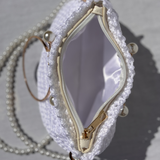 A close-up of a white textured woven clutch bag shown open, with a pearl chain strap, gold-tone zipper, and pearl embellishments on a light gray surface.